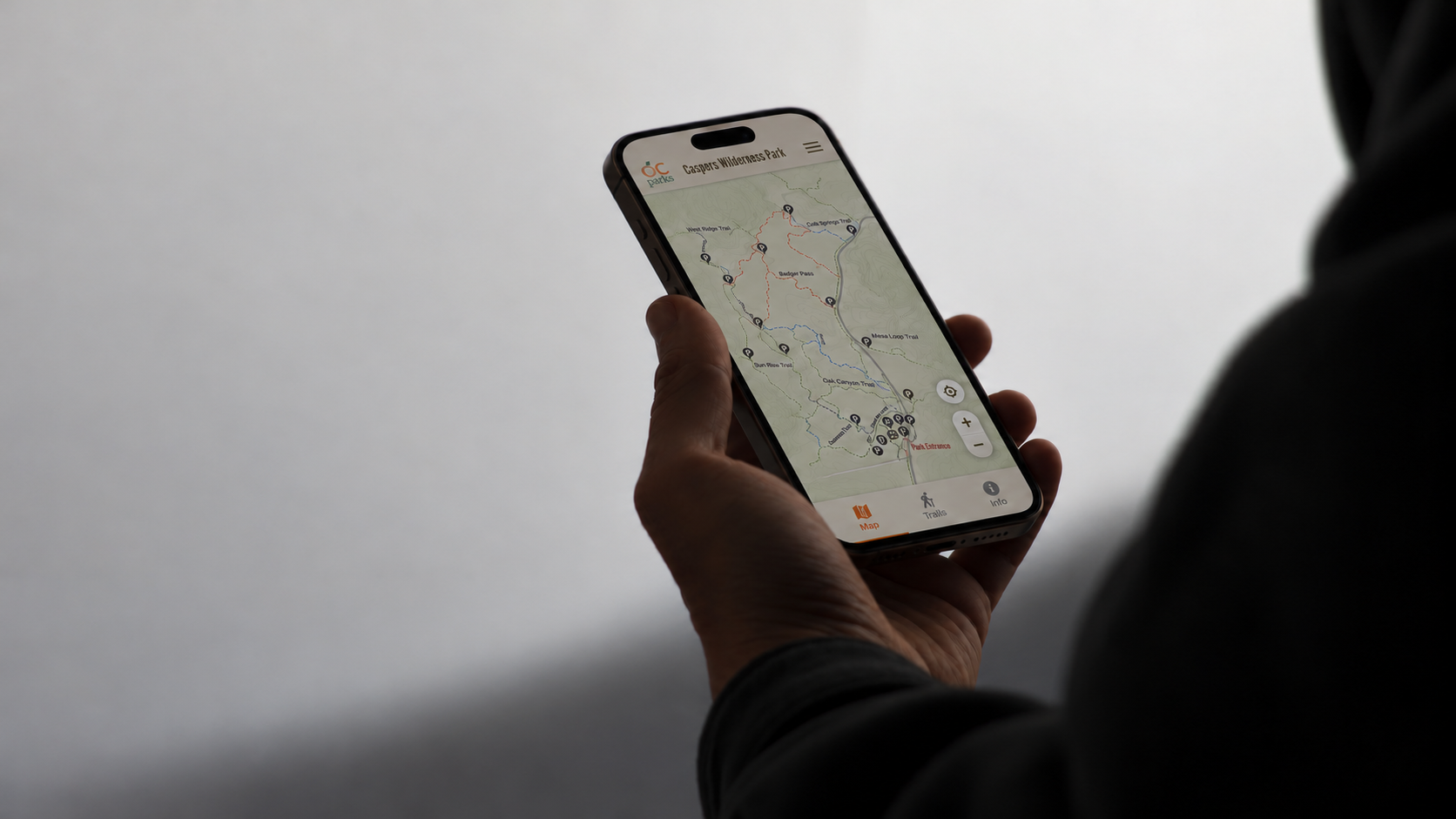 A visitor holds a smartphone showing the Caspers Wilderness Park trail map with OC Parks branding and Map, Trails, and Info navigation.