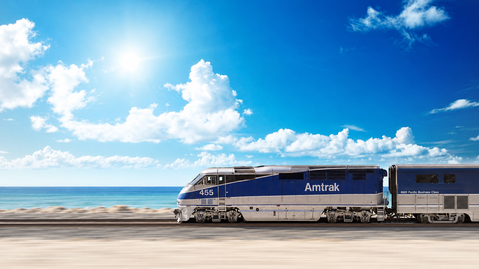 Amtrak locomotive 455 travels beside a turquoise ocean beneath a bright blue sky and billowing clouds, with motion-blurred tracks and foreground.