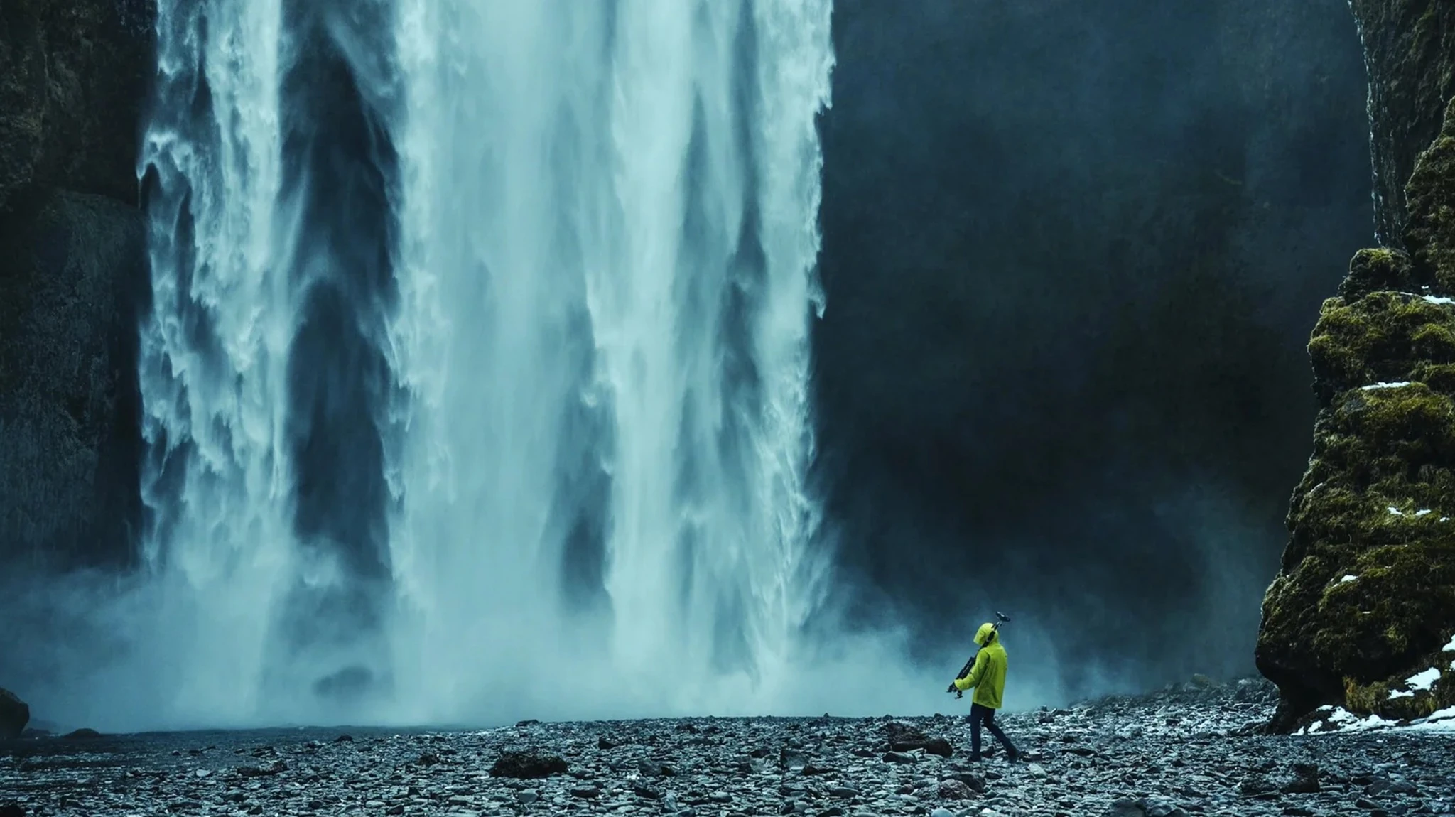 A person in a yellow jacket carries a camera tripod beside a large waterfall.