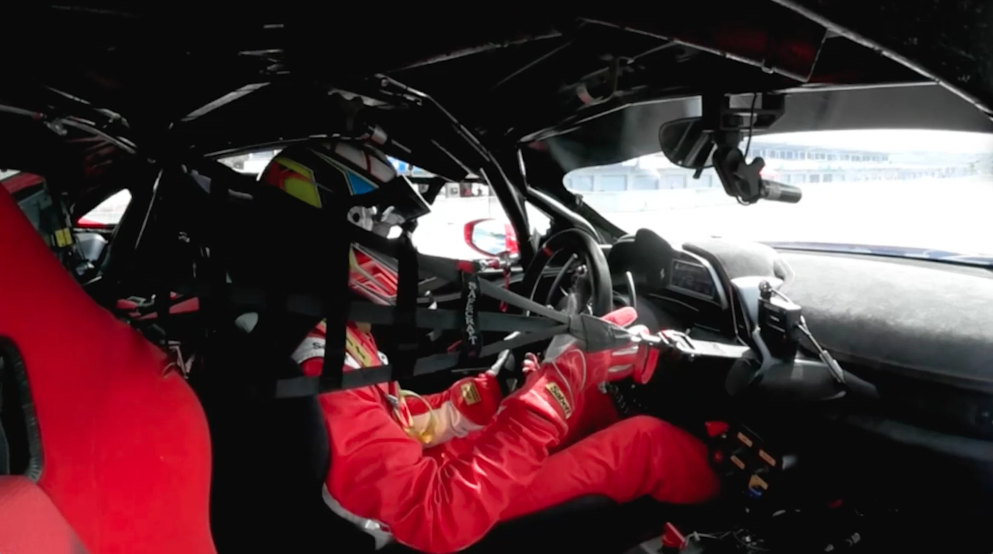 A helmeted driver in a red racing suit inside a race car, seen from the passenger side.
