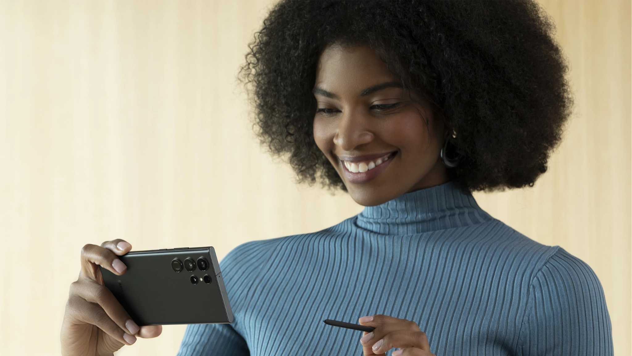 A woman holding a Samsung smartphone and S Pen against a warm beige background.